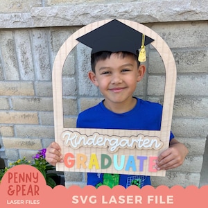 May include: A wooden graduation photo frame with the words "Kindergarten GRADUATE" in colorful letters. The frame has a black graduation cap on top and is held by a smiling child. The frame is a laser cut file.