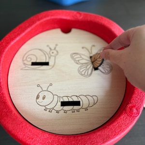 May include: A red plastic toy with a wooden insert featuring engraved illustrations of a snail, butterfly, and caterpillar. A hand is placing the butterfly piece into its slot. The toy is designed for children.