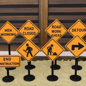 May include: A collection of miniature road signs in bright orange and black. The signs display messages like "Men Working," "Road Closed," and "Detour." Each sign is mounted on a black stand, creating a playful construction theme.