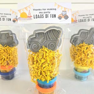 May include: A clear plastic party favor bag containing a gray plastic dump truck cookie cutter, yellow shredded paper filler, and a small container of modeling compound. The bag is sealed with a white card featuring orange bunting and the text "Thanks for making my party LOADS OF FUN" with a tractor illustration.