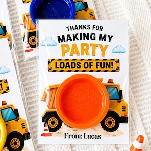 May include: A clear plastic favor bag containing a bright orange Play-Doh lid centered over a printed card featuring a yellow dump truck, blue clouds, and the text: THANKS FOR MAKING MY PARTY LOADS OF FUN! From: Lucas. A small orange traffic cone is nearby.