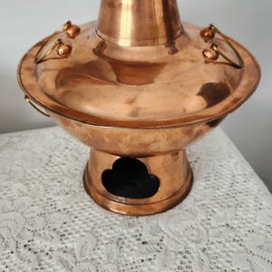 May include: A copper hot pot with a round base and a tall, cylindrical body. The pot has a lid with handles and a small chimney. The pot is sitting on a white lace tablecloth.