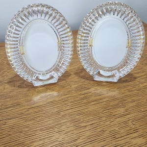May include: Two clear glass photo frames with a circular design. The frames have a ridged border and a small stand at the bottom. The frames are on a wooden surface.