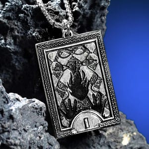May include: A rectangular silver-toned metal pendant necklace featuring an engraved tarot card design. The pendant displays a central flame motif, two hands reaching upward, an infinity symbol, and the Roman numeral I, all framed by a decorative border.