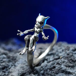 May include: A silver ring with a detailed sculpture of a creature with a humanoid form, pointed ears, and purple eyes. The ring is set against a dark blue background and a textured grey surface.
