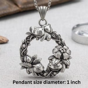 Peut inclure: Pendentif argenté représentant un cercle orné de trois Bulbasaur et de motifs floraux. Le pendentif mesure 2,5 cm de diamètre. Le collier présente une esthétique vintage et détaillée.