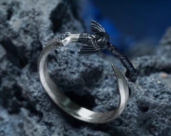 May include: A silver-toned ring with a black axe-shaped design. The ring features a curved band and a detailed axe head. The axe has a textured handle and a sharp blade. The ring is set against a dark, textured background.