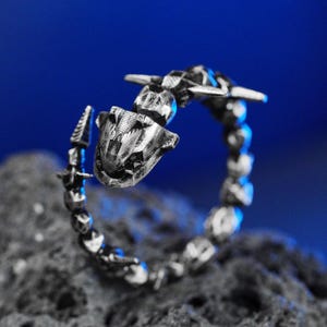 Op de afbeelding: Een zilverkleurige ring met een gedetailleerd, mechanisch ontwerp. De ring heeft een reeks onderling verbonden, afgeronde segmenten die de band vormen, met een prominent, gestileerd hoofdachtig element aan de bovenkant. De ring is geplaatst tegen een donkere, getextureerde ondergrond en een blauwe achtergrond.