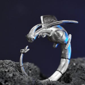Puede incluir: Un anillo de dragón plateado con detalles intrincados. El dragón está representado con alas y una cola curvada, frente a un objeto más pequeño y estilizado. El anillo está sobre un fondo oscuro y texturizado.