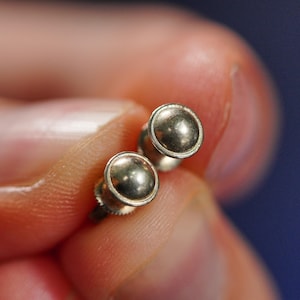 Puede incluir: Un par de pendientes de botón circulares de tono plateado con una superficie lisa y abovedada y un borde exterior elevado. Los pendientes tienen un acabado metálico y un diseño sencillo y redondeado, mostrando un aspecto pulido y brillante.