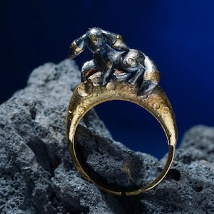 May include: A gold ring with a detailed rabbit design. The rabbit is dark grey with gold accents, sitting on a crescent-shaped band. The ring is set against a dark blue background and a textured grey surface.