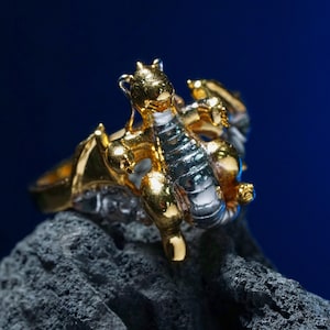 May include: A gold and silver dragon ring. The ring features a detailed dragon design with gold accents on the head, wings, and claws, and silver on the body. The ring is set against a dark blue background.