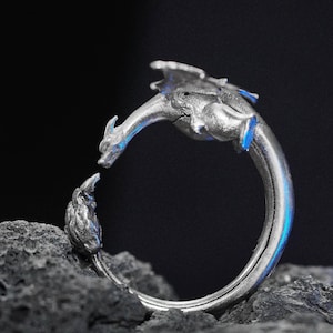 May include: A silver-colored dragon ring. The ring features a detailed dragon design with outstretched wings and a curved body. The dragon's head faces a flame-like shape. The ring is set against a dark background.