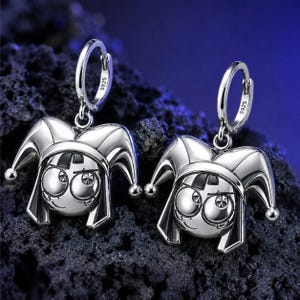 May include: A pair of silver hoop earrings featuring a jester head charm with a multi-pointed hat. Each hoop is stamped with S925. The jester face has large circular eyes and a small smile, with a polished metallic finish throughout the design.