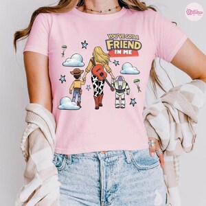 Op de afbeelding: Een lichtroze T-shirt met korte mouwen en een grafische print van een blond personage met een gitaar, een cowboy en een ruimteranger. De tekst luidt YOU'VE GOT A FRIEND IN ME. Het ontwerp bevat wolken, sterren en parachutespringende soldaten.