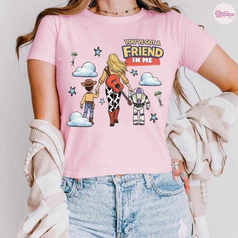 Op de afbeelding: Een lichtroze T-shirt met korte mouwen en een grafische print van een blond personage met een gitaar, een cowboy en een ruimteranger. De tekst luidt YOU'VE GOT A FRIEND IN ME. Het ontwerp bevat wolken, sterren en parachutespringende soldaten.
