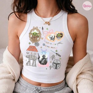 May include: White tank top featuring Star Wars-themed graphics, including Grogu, the Mandalorian, and the text "THIS IS THE WAY." The design incorporates various illustrations and text elements in a colorful arrangement.