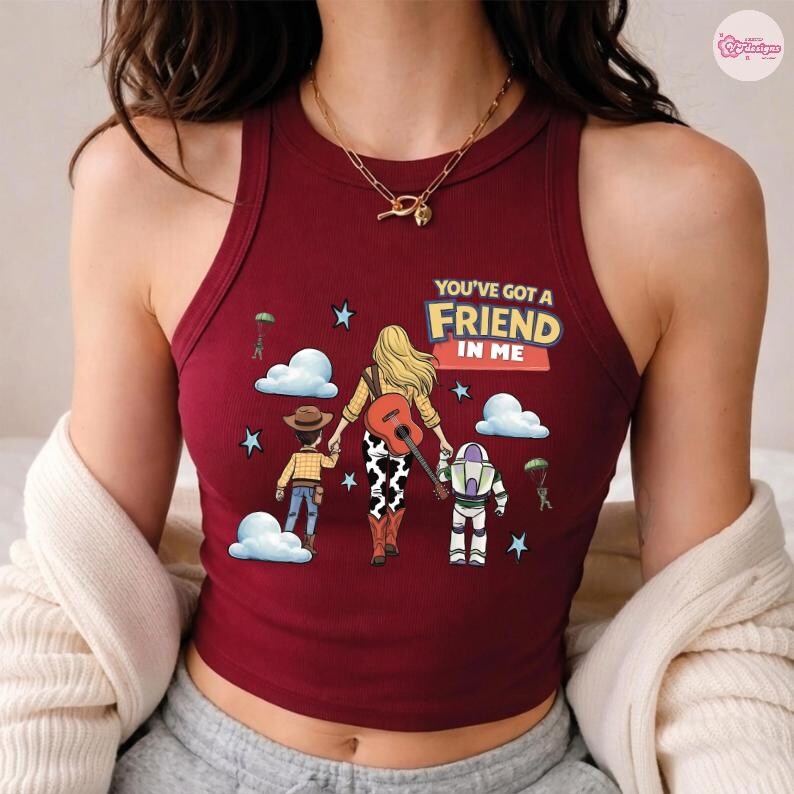 Op de afbeelding: Een bordeauxrood geribbelde tanktop met een grafische illustratie van een persoon met blond haar die een gitaar vasthoudt, geflankeerd door een cowboyfiguur en een space ranger-figuur. De tekst "YOU'VE GOT A FRIEND IN ME" staat erboven.
