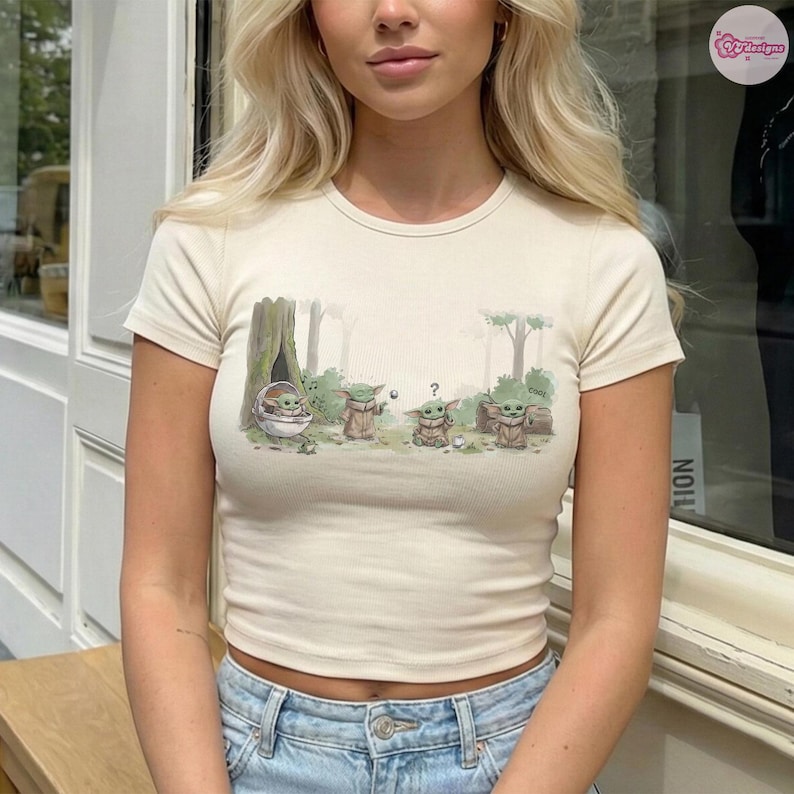 May include: Cream-colored, short-sleeved crop top featuring a cartoon illustration of Grogu from The Mandalorian. The graphic shows Grogu in a forest setting with other characters. The top is made of a ribbed fabric.