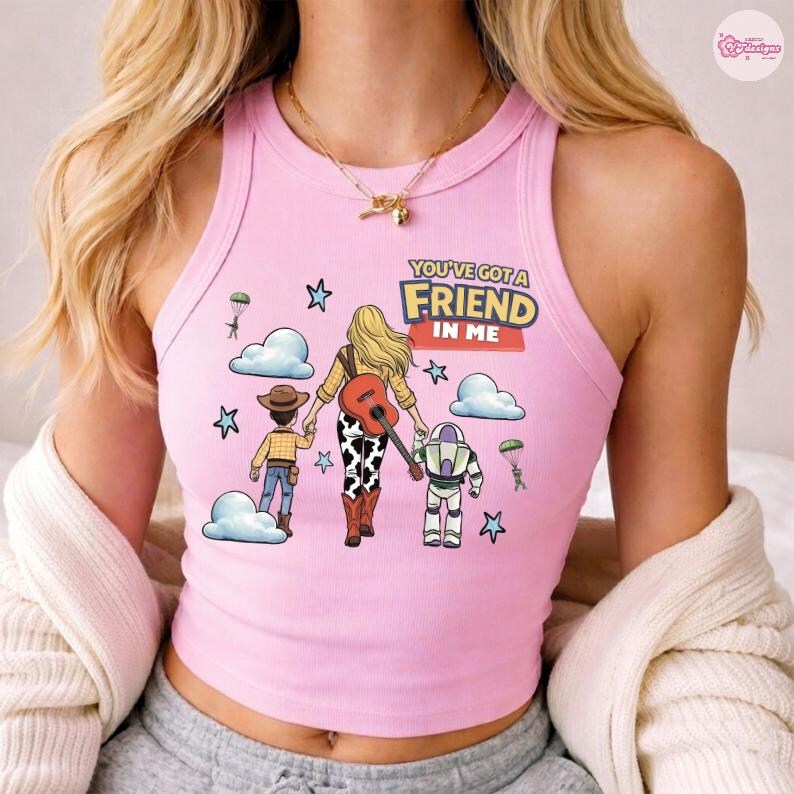 Op de afbeelding: Een lichtroze geribbelde tanktop met een grafische illustratie van een persoon met lang blond haar die een gitaar vasthoudt, vergezeld door een cowboyfiguur en een space ranger-figuur, met de tekst "YOU'VE GOT A FRIEND IN ME".