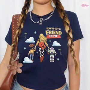 Op de afbeelding: Een marineblauw t-shirt met korte mouwen en een grafische print van drie wegwandelende figuren, met de tekst "YOU'VE GOT A FRIEND IN ME". Het ontwerp bevat wolken, sterren, parachutisten en een figuur die een gitaar op de rug draagt.