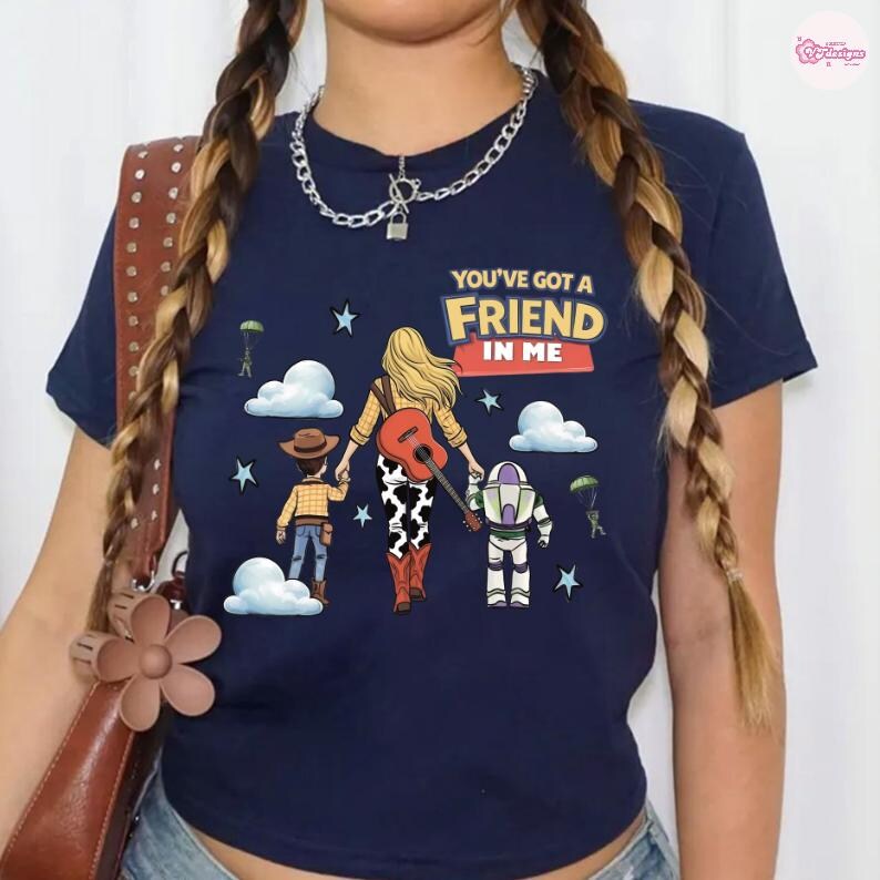 Op de afbeelding: Een marineblauw t-shirt met korte mouwen en een grafische print van drie wegwandelende figuren, met de tekst "YOU'VE GOT A FRIEND IN ME". Het ontwerp bevat wolken, sterren, parachutisten en een figuur die een gitaar op de rug draagt.