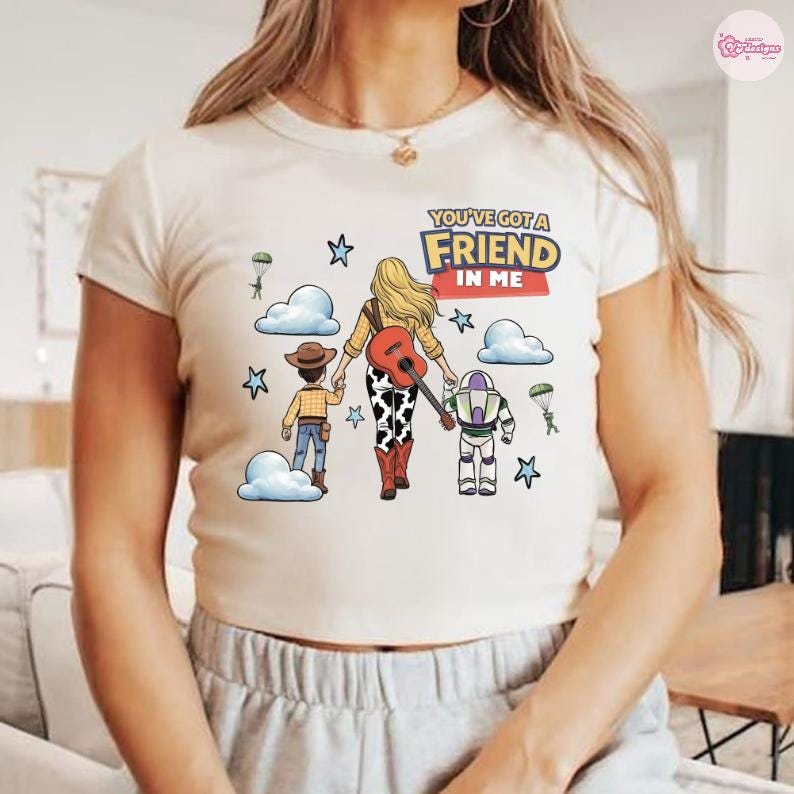 Op de afbeelding: Een cr&egrave;mekleurig T-shirt met korte mouwen en een afbeelding van een blond personage dat hand in hand loopt met een cowboy en een space ranger. De tekst luidt YOU'VE GOT A FRIEND IN ME. Het ontwerp bevat wolken, sterren en parachutes in cartoonstijl.