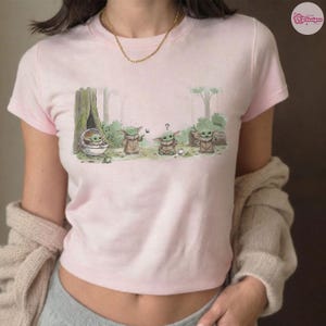 May include: Light pink t-shirt featuring a cartoon illustration of Grogu from The Mandalorian. The design shows Grogu in various poses in a forest setting. The word "cool" is written on the design.