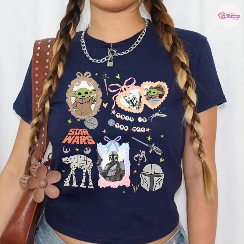 May include: Navy blue t-shirt featuring Star Wars graphics, including Grogu, the Mandalorian, and an AT-AT walker. The shirt has the words "Star Wars" and "This is the Way" printed on it. A chain necklace is worn.