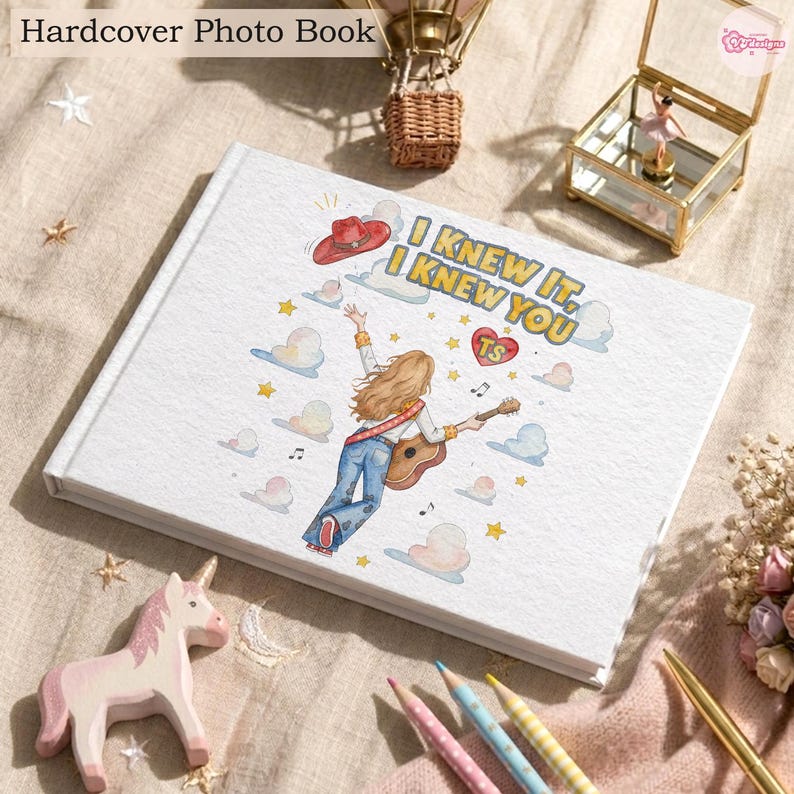 May include: A white hardcover photo book featuring a graphic illustration of a person with long hair playing an acoustic guitar, wearing blue jeans, and a red cowboy hat. The text reads I KNEW IT, I KNEW YOU with a red heart containing the letters TS.