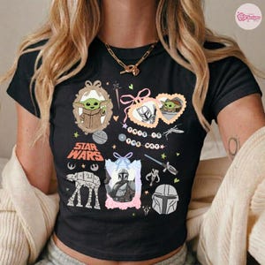 May include: Black t-shirt featuring Star Wars characters and imagery. The design includes illustrations of Grogu, The Mandalorian, and other elements from the Star Wars universe. The words "STAR WARS" are also printed on the shirt.