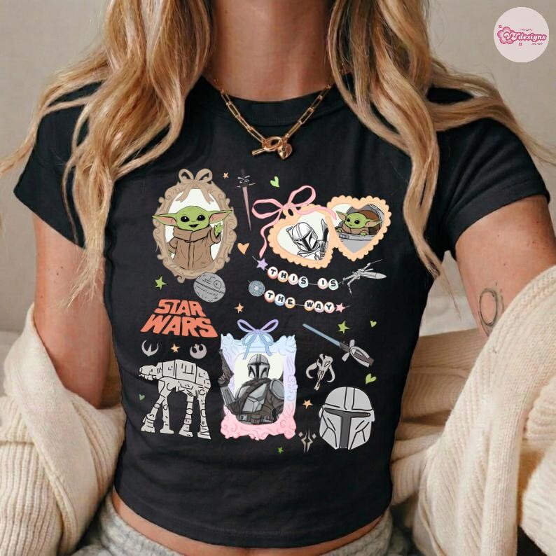 May include: Black t-shirt featuring Star Wars characters and imagery. The design includes illustrations of Grogu, The Mandalorian, and other elements from the Star Wars universe. The words "STAR WARS" are also printed on the shirt.