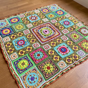 May include: A colorful crocheted blanket with a square pattern of flowers in various colors, including green, yellow, orange, red, pink, blue, and purple. The blanket has a crocheted border in orange, green, and white.