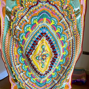Puede incluir: Una manta de ganchillo hecha a mano y de colores vibrantes. La manta presenta un diseño simétrico complejo con una forma de diamante central y patrones intrincados en tonos de amarillo, naranja, verde azulado, rosa y rojo. Los bordes están acabados con un borde rojo contrastante.