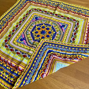 May include: A colorful crocheted blanket with a square design in the center. The blanket is made with a variety of colors, including yellow, blue, red, green, and pink. The blanket is folded in half and lying on a wooden floor.