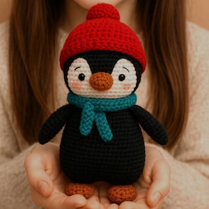 Op de afbeelding: Een handgemaakte gehaakte pinguïnpop met een rode gebreide muts en een turkooizen sjaal. De pinguïn heeft een zwart lichaam, een wit gezicht en een oranje snavel en voeten. De pop wordt in handen gehouden.