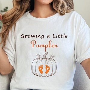 May include: White t-shirt with the text "Growing a Little Pumpkin" above a pumpkin graphic with baby footprints. The shirt is worn with blue jeans. The image is of a person sitting on a white couch.