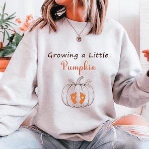 May include: A pale grey sweatshirt featuring the text "Growing a Little Pumpkin" and a pumpkin graphic with orange baby footprints. A comfortable, casual top, ideal for autumn or pregnancy announcements.