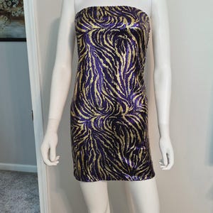 May include: A strapless mini dress featuring a vibrant sequin pattern in deep purple and metallic gold. The garment displays a swirling, abstract zebra-like design across the entire surface, creating a textured and reflective appearance throughout.