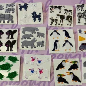 Puede incluir: Una colección de hojas de pegatinas con varios diseños de animales y vehículos. Las pegatinas incluyen rinocerontes, caniches, vacas, cebras, pingüinos, coches, tortugas, ovejas, tucanes y perros. Las pegatinas son de varios colores, incluyendo gris, negro, blanco, verde y rojo.