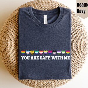 May include: Heather navy t-shirt with a row of colorful heart-shaped pride flags and the text "YOU ARE SAFE WITH ME". The shirt is displayed on a woven, circular surface.