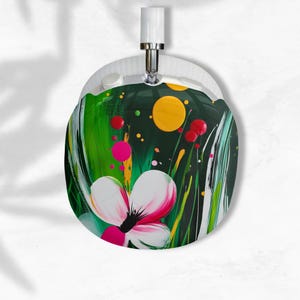 Könnte beinhalten: Runde, abstrakte Kunst mit einer weißen Blume mit rosa Akzenten vor einem dunkelgrünen Hintergrund mit gelben und roten Punkten. Grüne und gelbe Pinselstriche erzeugen einen grasartigen Effekt. Die Kunst ist an einer weißen, kreisförmigen Rückseite befestigt.
