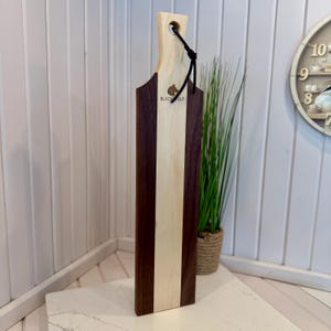 May include: A wooden serving board with a handle, featuring a vertical design of dark brown, light wood, and a central light wood stripe. The board has a hole in the handle and a black cord. The text "BLACK WOLF" and a wolf logo are visible.