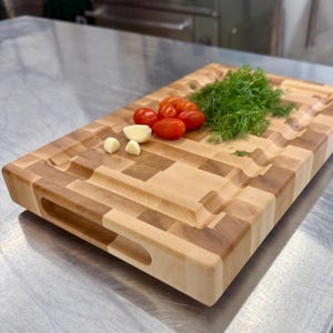 May include: A rectangular wooden chopping board with a light and dark wood pattern. The board features a carved juice groove and handle cutouts. Tomatoes, garlic, and dill are arranged on the board.