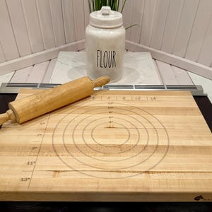May include: A wooden rolling pin lies on a light-coloured wooden cutting board with concentric circles and measurement markings. A white ceramic jar labelled "FLOUR" is in the background, suggesting a baking theme.
