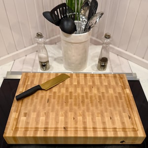 May include: A light-colored wooden butcher block with a rectangular shape and a carved groove around the edge. A gold-colored knife with a black handle rests on the block. Kitchen utensils are in a white ceramic container.