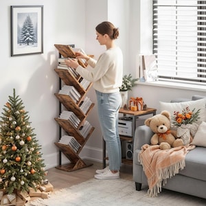May include: A brown and black tree-shaped bookshelf filled with books and CDs. A woman is standing next to the bookshelf. A small Christmas tree is in the foreground, and a couch with a teddy bear is in the background.