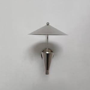 以下が含まれることがあります： シルバー仕上げのモダンなウォールスコンス。照明器具は円錐形のベースと三角形のシェードを備えています。スコンスは白い壁に取り付けられています。デザインはミニマリストで現代的です。