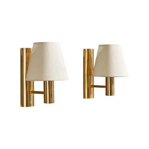 May include: A pair of wall sconces featuring polished gold-tone cylindrical metal backplates and arms. Each fixture supports a single off-white, tapered fabric lampshade. The design includes a vertical metal tube mount and a horizontal arm.