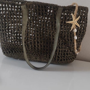 May include: Olive green woven tote bag with gray leather handles. A decorative jute rope with a starfish and small white beads hangs from the side. The bag is open at the top and sits on a white surface.